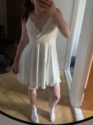 Vit spetsklänning med öppen rygg student!  - Säljer en vit kortklänning med spetsdetaljer och v-ringning perfekt som studentklänning !! . Klänningen har tunna axelband som korsas i ryggen och knyts, vilket ger en öppen rygg. Kjolen är lätt utsvängd och materialet känns som bomull eller polyester. Perfekt för sommarens alla tillfällen!