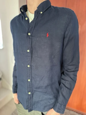 Ralph Lauren Linneskjorta – Marinblå (HELT NY) - Säljer en helt ny och oanvänd marinblå långärmad linneskjorta från Polo Ralph Lauren med röd logga. Det absolut favoritplagget för våren och sommaren. Skjortan är i nyskick och har aldrig blivit använd. • Märke: Polo Ralph Lauren • Material: 100% Linne • Storlek: Barn XL 18-20 (Motsvarar Small i herr) • Färg: Marinblå • Nypris: ca 1 900 kr • Skick: Helt ny / Nyskick 