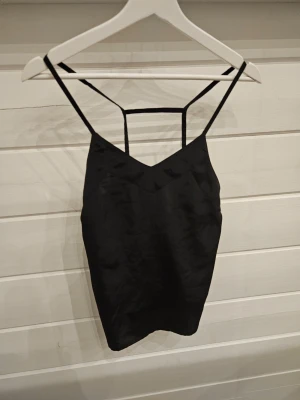 Svart linnetopp med tunna band - Snygg svart linnetopp med tunna axelband och unik detalj i ryggen. Toppen har en lätt glansig finish och är tillverkad i ett mjukt material som ger en skön känsla. Perfekt att styla med jeans eller kjol för en trendig look.