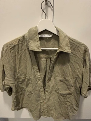 Beige croppad skjorta från Zara - Stilren beige skjorta från Zara med croppad passform och korta ärmar. Skjortan har två bröstfickor, klassisk krage och knäppning framtill. Perfekt för en avslappnad och trendig look.