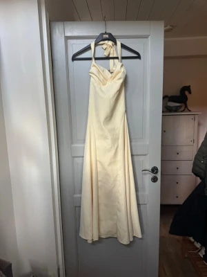 Lång ljus gul klänning med snörning - Elegant långklänning i ljus gulfärgad med halterneck och snygg snörning i ryggen. Klänningen har en mjuk, svepande form. Den är helt ny och ej använd. Perfekt för speciella tillfällen.