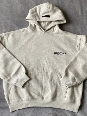 Essentials Fear of God hoodie - Snygg ljusgrå hoodie från Essentials Fear of God med svart logga på bröstet och stor text på ryggen. Klassisk känguruficka, ribbade muddar och en rejäl huva. Perfekt för dig som gillar streetwear och vill ha en clean look.