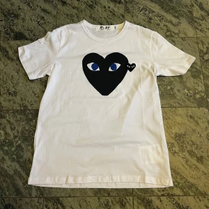 Vit Comme des Garçons Play t-shirt - Vit t-shirt från Comme des Garçons Play med det ikoniska svarta hjärtat med ögon tryckt på bröstet. Klassisk passform, rund hals och korta ärmar. Tillverkad i mjuk bomull, perfekt för en clean och stilren look. Skulle säga att den är lite mer passform för folk som har S!