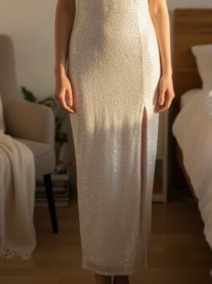 Glittrig långklänning med slits - Elegant glittrig långklänning i ljus ton med smal passform och hög slits framtill. Klänningen har ärmlös design och skimrar vackert i ljuset, perfekt för speciella tillfällen. Materialet känns mjukt och följsamt mot kroppen.