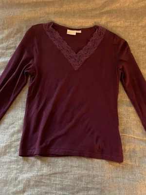 Vinröd topp med spetsdetalj - Vinröd långärmad topp med v-ringning och dekorativ spets runt halsen. Tillverkad i mjukt material som känns skönt mot huden. Perfekt för dig som gillar stilrena plagg med en liten twist.