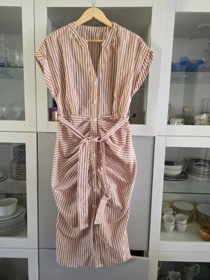 Randig beige/brun och vit klänning med knyt - Säljer en snygg randig klänning i beige och vitt med knappar framtill och ett matchande knytskärp i midjan. Klänningen har korta ärmar och siluettform, perfekt för varma dagar. Tillverkad i linne som känns skön mot huden. Ingen stretch och ganska tight, mer M/L än L/XL