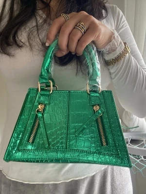 Grön metallic handväska med krokoprägling - Säljer en grön metallic handväska med krokodilpräglat mönster och guldfärgade detaljer. Väskan har två handtag, två dekorativa dragkedjor framtill och en fyrkantig form. Perfekt statement piece för dig som vill sticka ut.
