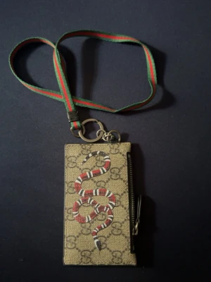  plånbok med orm och halsband - Snygg plånbok från  med klassiskt GG-mönster och en röd, vit och svart orm på framsidan. Beige bakgrund och grön-röd halsrem. Plånboken har dragkedja på sidan och metallringar för nycklar eller accessoarer. Perfekt för dig som gillar lyxiga detaljer. (Den är äkta)