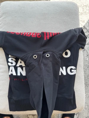 Svart hoodie Helmut Lang - Cool svart hoodie från Helmut Lang med stort vitt och rött texttryck på ryggen. Klassisk huva med snörhål och ribbade muddar. Limiterad.