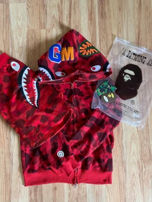 Röd BAPE Shark Hoodie med kamouflage - Säljer en ikonisk röd BAPE Shark Hoodie med kamouflagemönster och hajmotiv på huvan och ärmarna. Dragkedja framtill, broderade detaljer och färgglada bokstäver på huvan. Tillverkad i mjuk bomull, perfekt för streetwear och hypebeasts.