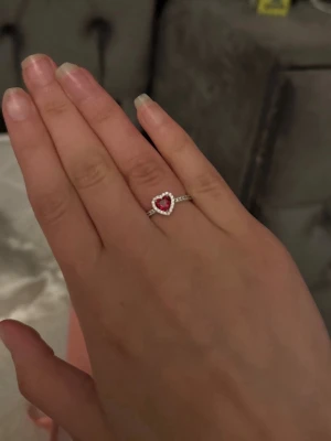 Silverring med hjärtformad röd sten - Snygg silverfärgad ring med en hjärtformad röd sten i mitten, omgiven av små klara stenar längs kanten och bandet. Perfekt för dig som gillar romantiska detaljer och vill ha något som sticker ut lite extra.