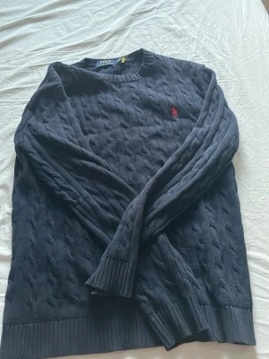 Mörkblå kabelstickad tröja Polo Ralph Lauren - Mörkblå kabelstickad tröja från Polo Ralph Lauren med klassisk röd broderad logga på bröstet. Tröjan har rund halsringning, långa ärmar och ribbade muddar. Perfekt för en stilren och avslappnad look.