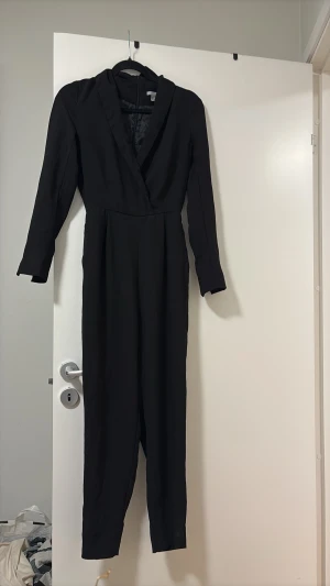 Svart långärmad jumpsuit med krage - Snygg svart jumpsuit med långa ärmar och djup omlott-ringning. Har en markerad midja och smala ben. Klassisk krage ger en stilren look. Perfekt för dig som vill ha en enkel men trendig outfit.