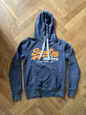 Blå Superdry hoodie med tryck - Mörkblå hoodie från Superdry med stor orange och vit logga på bröstet samt japansk text. Klassisk känguruficka framtill, vita dragsnören i huvan och ribbade muddar. Mjuk bomullsblandning som känns skön mot huden. Perfekt för en avslappnad streetwear-look.
