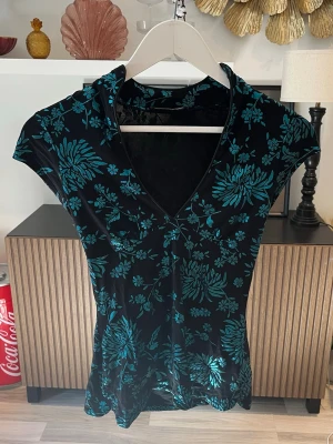 Svart blommig topp med v-ringning - Cool topp i svart med turkost blommigt mönster och v-ringad hals från Jane norman. Kortärmad och figurnära passform, perfekt för att ge din outfit en unik touch. Materialet är mjukt och mönstret är glansigt, vilket gör den extra bekväm och snygg. Står ingen storlek men skulle säga S. Axelbredd: 45cm. Längd: 62cm. Byst: 40cm.