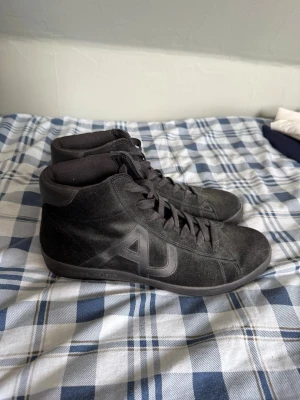 Svarta Armani Jeans sneakers - Snygga svarta sneakers från Armani Jeans med AJ-logga på sidan. Skorna har hög modell, snörning och är tillverkade i mockaliknande material med textildetaljer. Perfekta för dig som gillar stilrena och klassiska sneakers.