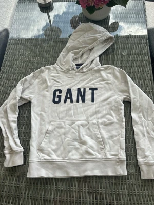 Vit hoodie från GANT  - Snygg vit hoodie från GANT med stor mörkblå logga på bröstet. Stl 146/152. Trycket är lite slitet men inget man tänker så mycket på. Det finns en liten fläk på vänster arm (se bild). 