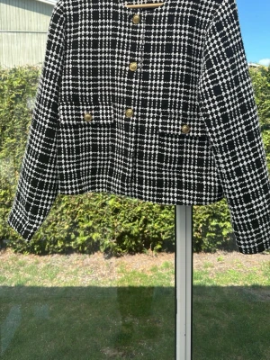 Svartvit rutig kavaj First Class - Stilren svartvit rutig kavaj från First Class Collection med guldknappar och två fickor framtill. Kavajen har rund halsringning och är tillverkad i ett strukturerat tyg som ger en lyxig känsla. Perfekt för dig som vill sticka ut med en klassisk men trendig look.
