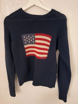 Mörkblå stickad tröja Polo Ralph Lauren - Mörkblå stickad tröja från Polo Ralph Lauren med amerikansk flagga i rött, vitt och blått på bröstet. Tröjan har rund hals, långa ärmar och ribbstickade muddar. Perfekt för dig som gillar klassisk och cool stil.