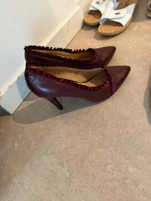 Vinröda pumps med volangkant - Säljer ett par vinröda pumps från MISHOO med spetsig tå och dekorativ volangkant i satin runt öppningen. Skorna har smal klack och är tillverkade i ett glansigt material som ger en elegant look. Perfekta för dig som vill sticka ut lite extra.