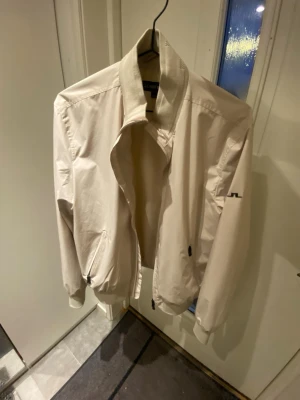Regnjacka från J.Lindeberg - Stilren beige regnjacka från J.Lindeberg med dragkedja, ribbade muddar och diskret logga på ärmen. Jackan har två fickor framtill och är tillverkad i ett lätt, slätt material som passar perfekt till vår, höst och regniga sommardagar.   Nypris: 3200 kr. Köpte jackan för ett år sedan men har endast använt den ett fåtal gånger, så den är typ nyskick. Hör gärna av dig vid några funderingar eller fler bilder! (Priset är inte hugget i sten) 😉😊