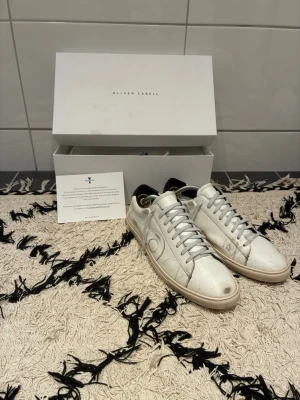 Oliver Cabell skor - Snygga vita sneakers från Oliver Cabell i klassisk låg modell. Skorna är tillverkade i slätt läder med vita snören och diskret logga på sidan. Yttersulan är ljus och kontrasterar snyggt mot ovandelen. Perfekta för en clean och stilren look.