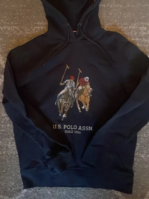 Svart hoodie från U.S. Polo Assn. - Snygg svart hoodie från U.S. Polo Assn. med stort broderat motiv av två polospelare på hästar framtill. Klassisk huva med dragsko och ribbade muddar vid ärmar och nederkant. Perfekt för dig som gillar sportig och stilren streetwear.
