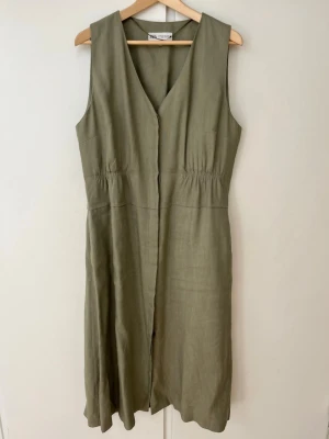 Olivgrön ärmlös klänning från Zara - Olivgrön klänning från Zara i linne med v-ringning och ärmlös design. Klänningen har en markerad midja med lätt rynk och en rak, avslappnad passform med knappar. Perfekt för varma dagar och enkel att styla.