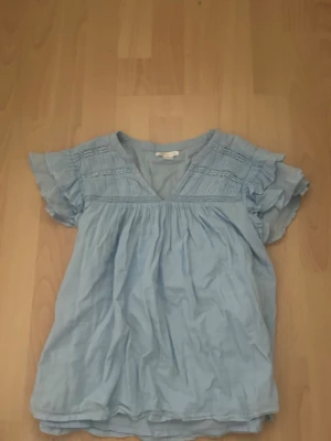 Ljusblå volangblus - Supersöt ljusblå blus från Gina tricot med volangärmar och v-ringning. Blusen har fina spetsdetaljer upptill. Den är i barnstorlek 158/164