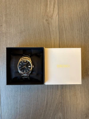 Seiko klocka med svart urtavla - Stilren Seiko klocka med svart urtavla och datumvisning. Klockan har ett klassiskt armband i silverfärgad metall och tydliga indexmarkeringar. Perfekt accessoar för dig som gillar en clean och tidlös look. Helt ny och man får men allt, kvitto, extra länkar, box osv. Klockans nypris är 3600 men mitt pris är 1999! Skriv vid minsta fundering!