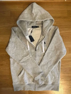 Grå zip hoodie från Polo Ralph Lauren - Snygg gråmelerad hoodie med dragkedja från Polo Ralph Lauren. Klassisk huva med vita snören, två fickor framtill och den ikoniska mörkblå loggan broderad på bröstet. Tillverkad i mjukt bomullsmaterial för en skön och avslappnad look.