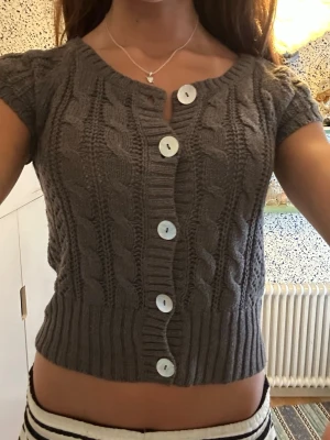 Grå kabelstickad kortärmad kofta - Supersöt grå kabelstickad kofta från L.L.Bean med korta ärmar och stora vita knappar framtill. Croppad modell med bred ribbad kant nedtill och runt ärmslut. Perfekt att styla över ett linne eller t-shirt för en trendig look.