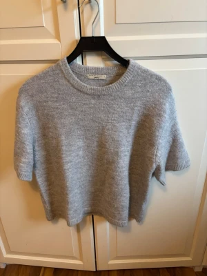 Grå stickad tröja Gina Tricot - Mjuk och fluffig grå stickad tröja från Gina Tricot med korta ärmar och rund hals. Tröjan har en enkel, rak passform och ribbstickade detaljer vid hals, ärmslut och nederkant. Perfekt för lager på lager och en stilren look.