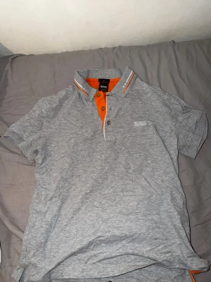 Hugo boss piké  - Snygg grå pikétröja från BOSS med orange insida vid knäppningen och orange/vita ränder på kragen. Tröjan har korta ärmar, klassisk krage och knappar framtill. Perfekt för en stilren och sportig look.