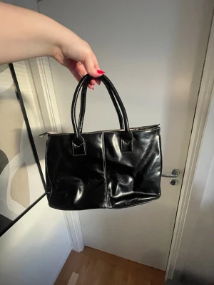 Svart handväska i skinnimitation - Stilren svart handväska i skinnimitation med två handtag och kontrastsömmar. Perfekt storlek för vardagens essentials och enkel att matcha med olika outfits