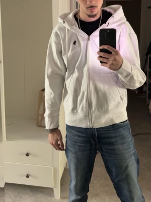 Vit polo Ralph zip up hoodie  - Säljer en vit hoodie med dragkedja från Polo Ralph Lauren. Hoodien har huva med snören, två fickor framtill och den klassiska lilla loggan broderad på bröstet. Perfekt för en avslappnad och stilren look.