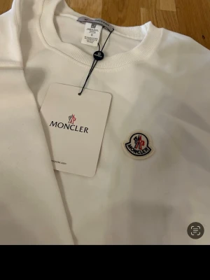 Vit sweatshirt från Moncler - Snygg vit sweatshirt från Moncler med rund halsringning och ribbade muddar. Tröjan har Monclers ikoniska logotyp broderad på bröstet och en svart detalj på ena ärmen. Perfekt för en clean och stilren look.
