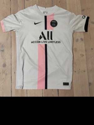 PSG matchtröja vit/rosa Nike - Snygg PSG matchtröja i vitt med en rosa och svart vertikal rand på framsidan. Tröjan har korta ärmar, svart Nike-logga och PSG-emblem på bröstet samt sponsortryck. Ena ärmen är svart och den andra rosa. Tillverkad i lätt och ventilerande material.