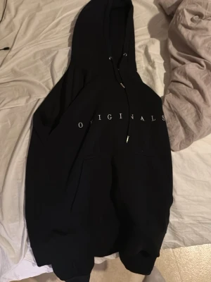 Den svarta originala hoodien från Jack - Jones  - Svart hoodie från Jack Jones med stor huva och dragsko. Framtill finns ett diskret vitt tryck med texten 'ORIGINAL ' över bröstet och en klassisk känguruficka. Perfekt för en avslappnad och stilren look.