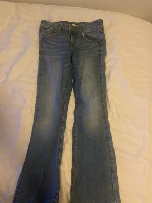 Blå bootcut jeans  - Säljer ett par blå bootcut jeans med lätt slitna detaljer och klassisk femficksmodell. Jeansen har en snygg tvätt och är tillverkade i denim med lite stretch för extra komfort. Perfekta för en avslappnad och trendig look.