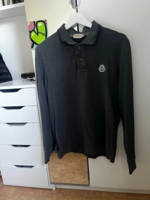 Moncler Pike Långärmad - Säljer en mörkgrå långärmad skjorta från Moncler med klassisk krage och knappslå framtill. Skjortan har Moncler-logga på bröstet och är tillverkad i mjukt bomullsmaterial. Perfekt för en clean och stilren look.
