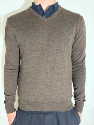 Brun stickad tröja från Ralph Lauren - Snygg brun stickad tröja från Ralph Lauren med diskret grönt logobroderi på bröstet. Tröjan har V-ringning, långa ärmar och ribbade muddar vid ärmslut och nederkant. Perfekt att bära över en skjorta för en clean och stilren look. Storlek L men passar även M