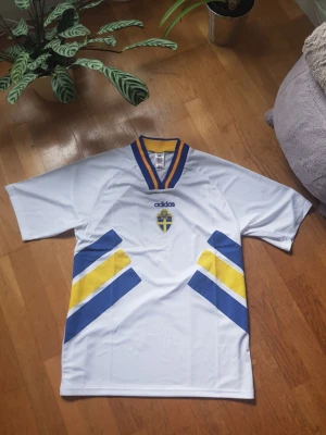 Svensk landslagströja Adidas XL - Vit svensk landslagströja från Adidas med blå och gula detaljer samt broderad svensk sköld på bröstet. Tröjan har V-ringad krage med blå och gula ränder och är tillverkad i ett lätt och ventilerande material, perfekt för fotboll eller träning.