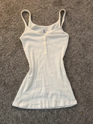 Vit ribbad linnetopp med knappar - Snygg vit ribbad linnetopp med smala axelband och knappar framtill från brandy Melville! Storleken är one size men skulle säga att den passar som en S! Säljer då den är lite för lång på mig, knappt använd