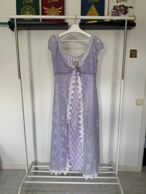 Ljuslila långklänning från Lacemade - Långklänning i ljuslila med vita spetsdetaljer längs kanter och ärmar samt en empire waist. Klänningen kan knytas i ryggen för att framhäva kurvor! Här är måtten från LaceMades hemsida! Bröst: 109cm, Midja: 98cm