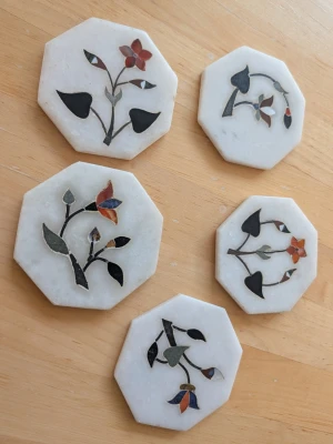 Sexkantiga marmorunderlägg med blommor - Fem stycken sexkantiga underlägg i vit marmor med färgglada blommönster inlagda i sten. Varje underlägg har unika blommor i rött, svart, grönt och blått. Perfekta som dekoration eller för att ställa glas på. Säljes som set.