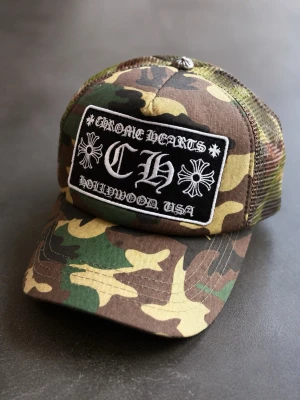 Chrome Hearts camo keps - Säljer en grym keps från Chrome Hearts med camo-mönster och mesh på sidorna. Stor broderad patch framtill med logga och texten 'Hollywood USA'. Perfekt för dig som vill sticka ut med streetstyle och exklusiv känsla.