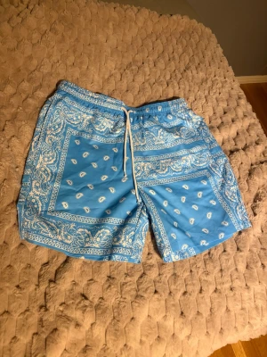 Blå bandanamönstrade shorts med snörning - Snygga blå shorts med vitt bandanamönster och snörning i midjan. De har elastisk midja och avslappnad passform, perfekta för sommardagar. Materialet känns lätt och bekvämt, och shortsen har fickor både fram och bak.