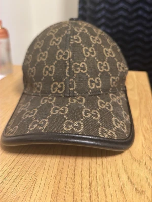 Gucci keps med GG-mönster brun - Snygg brun keps från Gucci med klassiskt GG-monogram i beige över hela kepsen. Bågen är böjd och har en svart läderdetalj längs kanten samt justerbar rem bak. Perfekt accessoar för att lyfta din streetwear-look.