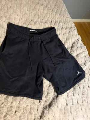 Svarta Jordan shorts med snörning - Säljer ett par svarta shorts från Jordan med klassisk logga broderad i vitt på vänster ben. Shortsen har snörning i midjan och är tillverkade i mjuk bomull, perfekta för en avslappnad och sportig stil.
